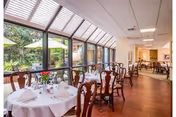 Dining room with round tables covered in white tablecloths, set with glassware, plates, and napkins. The room has large windows and skylights allowing natural light to fill the space, with wooden chairs around the tables and a view of greenery outside.