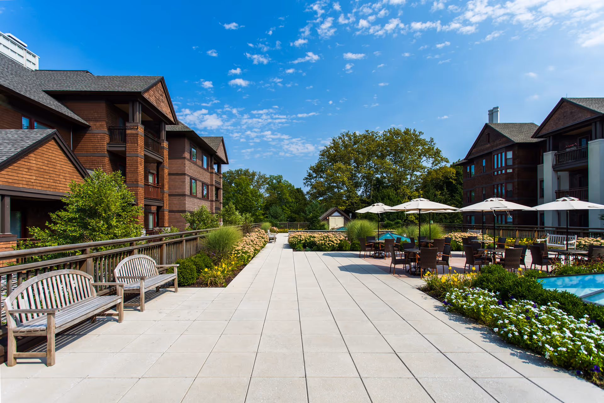 Outdoor patio area at Edgehill Senior Living Community Stamford with wooden benches along a railing on the left, tables with umbrellas and chairs on the right, surrounded by greenery and flowers, with multi-story residential buildings in the background under a blue sky with scattered clouds.
