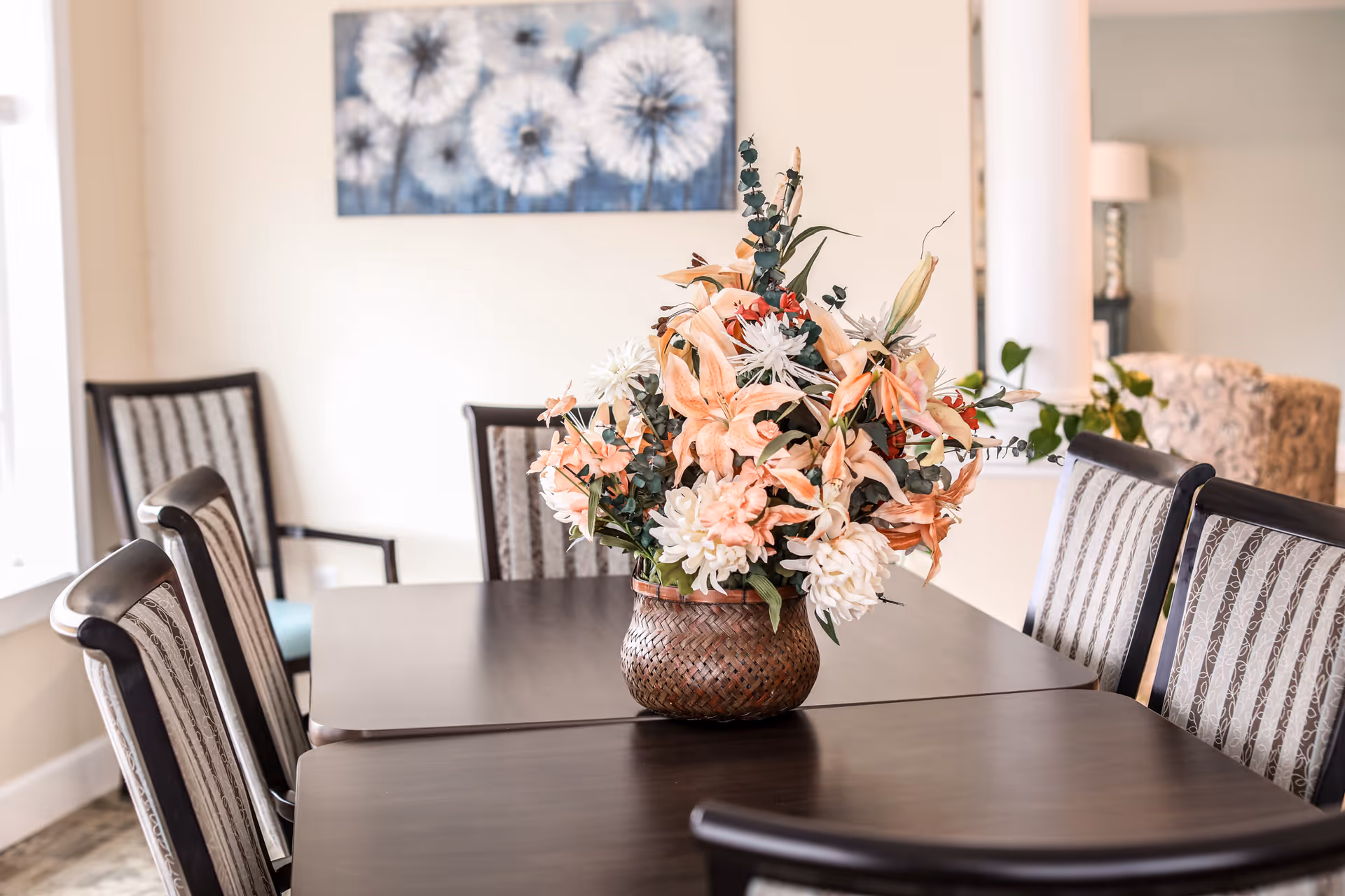 A dining table with a woven basket centerpiece filled with a variety of artificial flowers in soft peach and white colors. The table is surrounded by six upholstered chairs with patterned fabric. In the background, there is a wall with a painting of dandelions and part of a living area with a lamp and a patterned armchair.