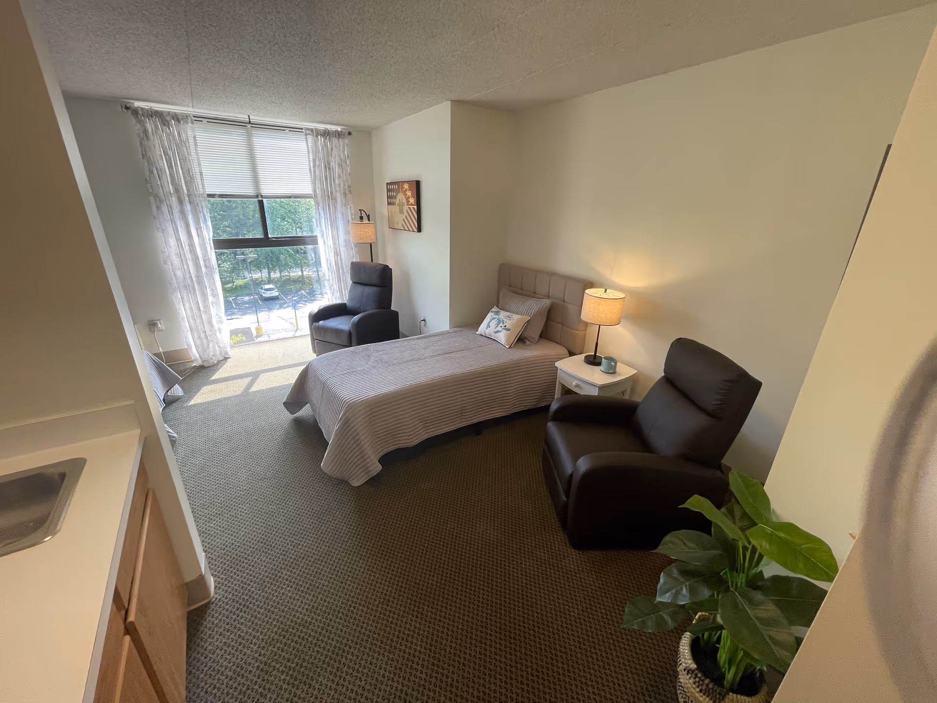 A small, cozy bedroom in a senior living facility with a single bed covered in a striped bedspread, two dark reclining chairs, a white nightstand with a lamp and a mug, a large window with sheer curtains letting in natural light, and a potted plant in the corner.