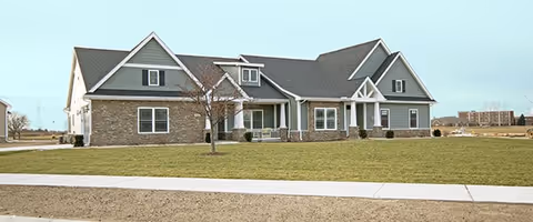 Exterior view of a single-story residential-style building with gray siding and stone accents, multiple gables, and a covered front porch, surrounded by a large grassy lawn and a sidewalk in front.