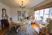 A bright dining room with a round white table surrounded by six chairs with floral cushions. The room features a large window with floral valance, two striped armchairs near the window, a chandelier hanging from the ceiling, a wooden side table with a mirror above it, and a patterned area rug on the wooden floor.