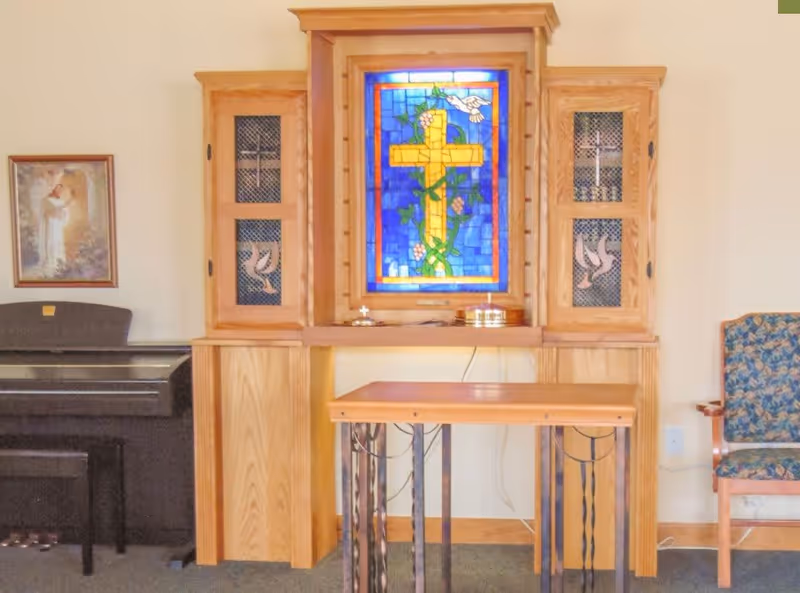 A small chapel or prayer room with a wooden altar featuring a stained glass window depicting a yellow cross with green vines, pink flowers, and a white dove on a blue background. On either side of the altar are wooden cabinets with mesh doors displaying crosses and doves. To the left is a black piano with a framed religious picture above it, and to the right is a wooden chair with a floral patterned cushion.