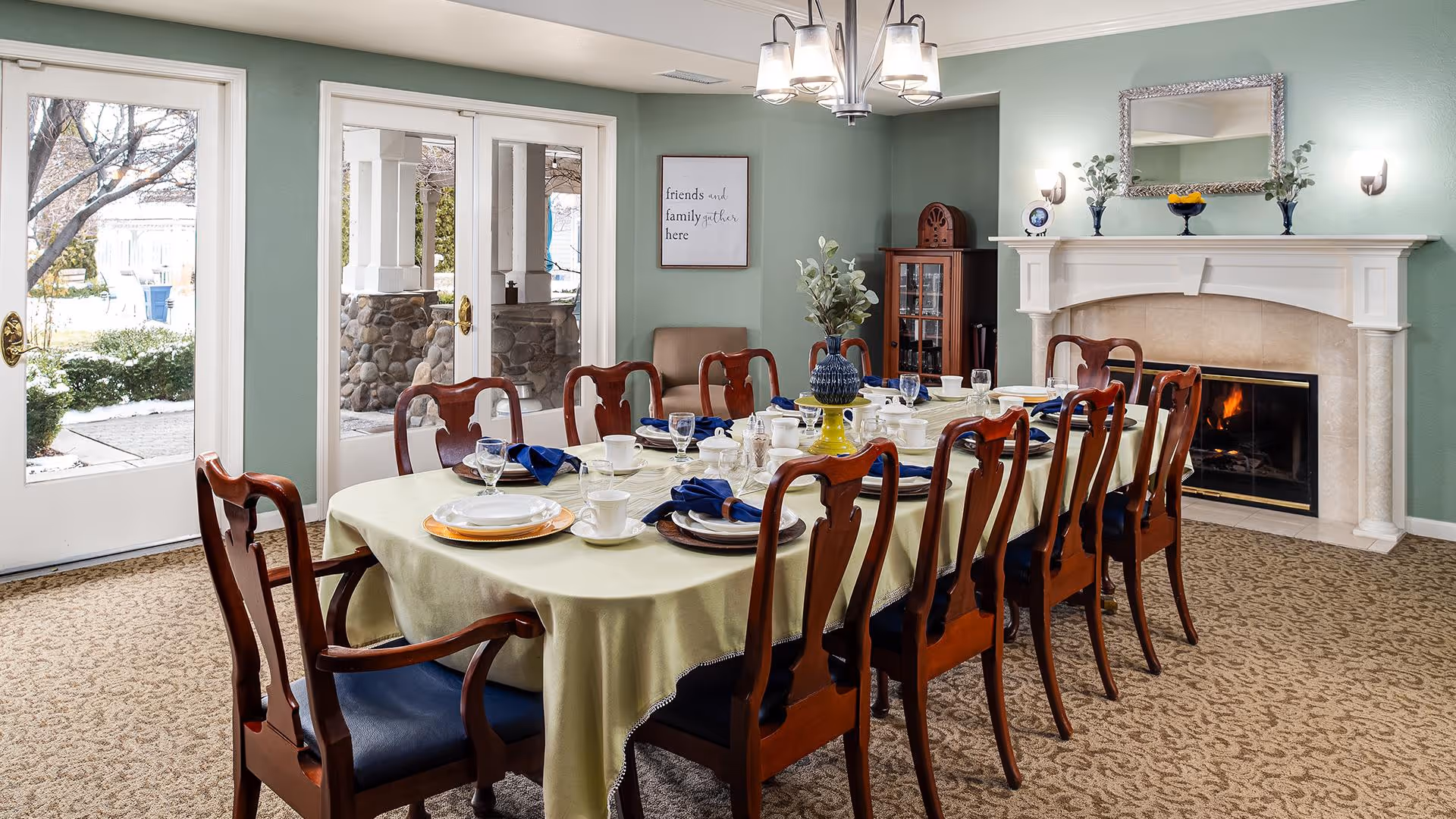 A formal dining room with a long table covered with a light green tablecloth, set with plates, cups, glasses, and blue napkins. The room has wooden chairs with blue cushions around the table. There is a fireplace with a fire burning on the right side, decorated with a mirror, vases, and a bowl of fruit. A framed sign on the wall reads 'friends and family gather here'. Glass doors on the left open to an outdoor area with trees and shrubs.