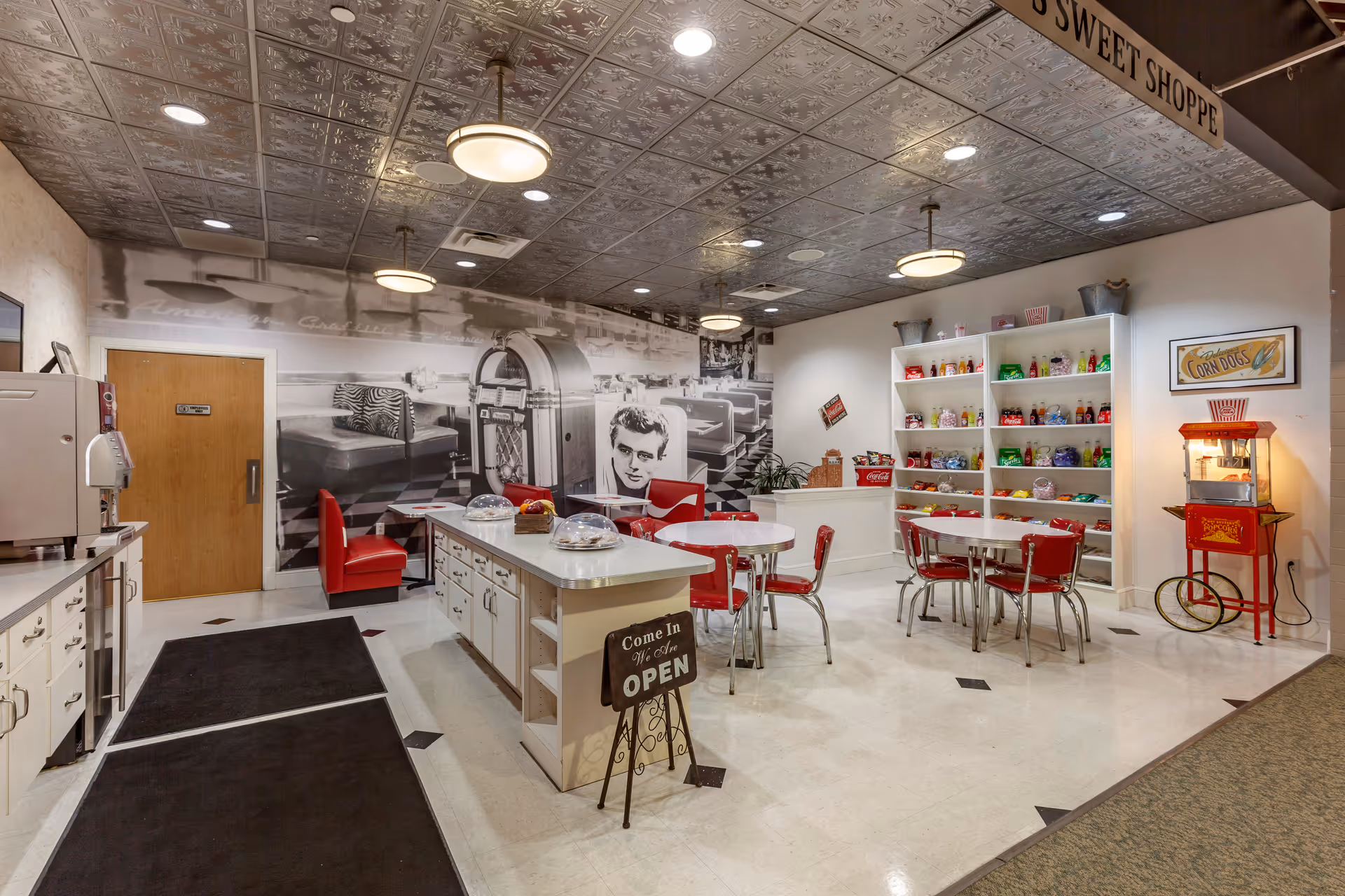 Interior of a retro-style sweet shop with red chairs and tables, a popcorn machine, shelves stocked with colorful candy and soda bottles, a black and white mural of a vintage diner scene on the wall, and a sign that says 'Come In We Are OPEN' on a small stand near the center island.