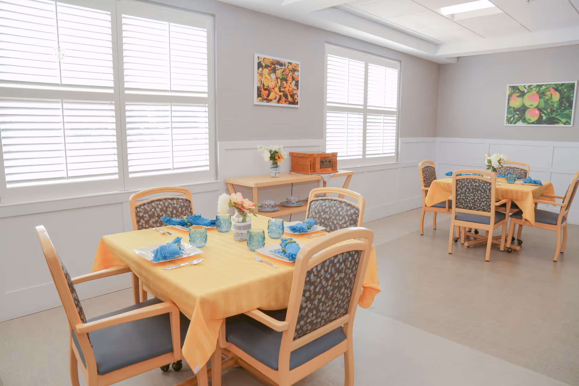 Bright dining room with tables set with yellow tablecloths, blue napkins, and patterned chairs near shuttered windows.