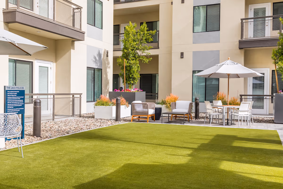 Outdoor courtyard area at Overture Kierland featuring a green lawn, modern patio chairs, tables with umbrellas, potted plants, and a multi-story building with balconies and windows in the background.
