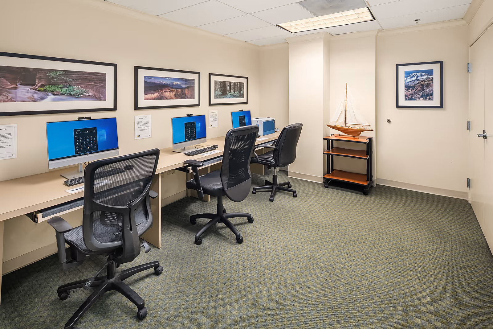 A small computer room with three desktop computers on a long desk, each accompanied by a black office chair. The walls are decorated with framed landscape photographs. There is a model sailboat on a small wooden cart in the corner of the room. The floor is carpeted in a green pattern and the ceiling has fluorescent lighting.