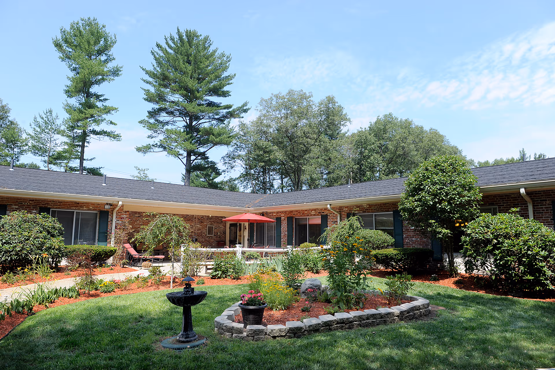 A sunny outdoor courtyard at Sudbury Pines Extended Care featuring a well-maintained garden with various plants and flowers, a small black birdbath, and a red umbrella shading a seating area. The courtyard is surrounded by a single-story brick building with windows and greenery, under a blue sky with some clouds.