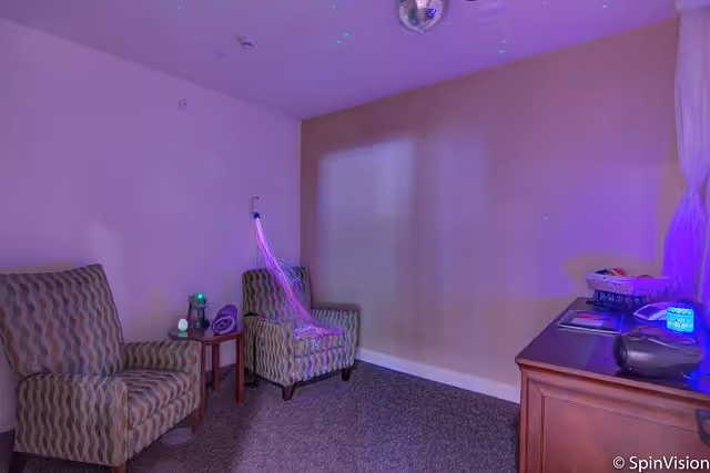 Small softly lit sitting room with two patterned armchairs, a side table with glowing sensory lights, a cabinet with devices, and a disco ball on the ceiling.