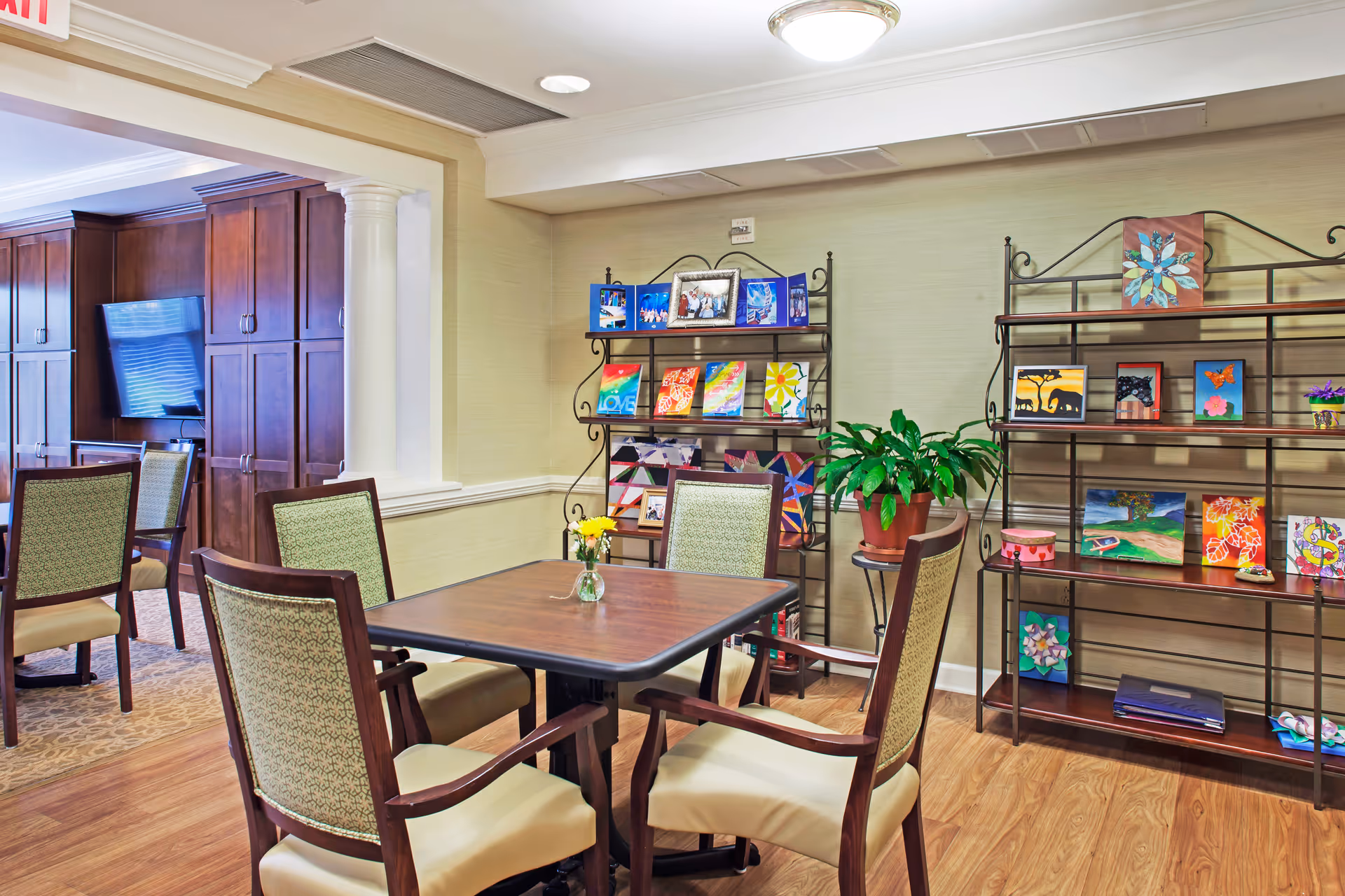 A cozy common area with a wooden table and four cushioned chairs. Behind the table are two metal shelves displaying colorful artwork and a potted plant. In the background, there is a wall-mounted TV and wooden cabinetry.