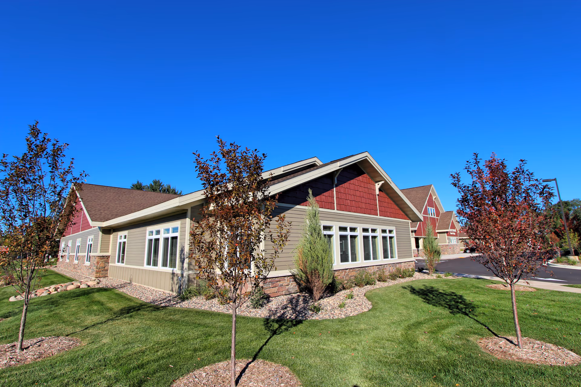 Exterior view of a single-story building with beige and red siding, multiple windows, and a brown roof. The building is surrounded by a well-maintained lawn with small trees and landscaping rocks under a clear blue sky.