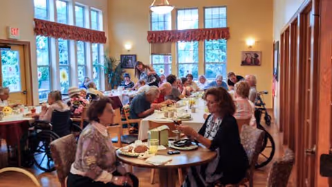 A dining room in a senior living facility with several elderly residents seated at round tables, eating and socializing. The room has large windows with red valances, warm yellow walls, and wooden flooring. Some residents are in wheelchairs, and a caregiver is assisting one of them.