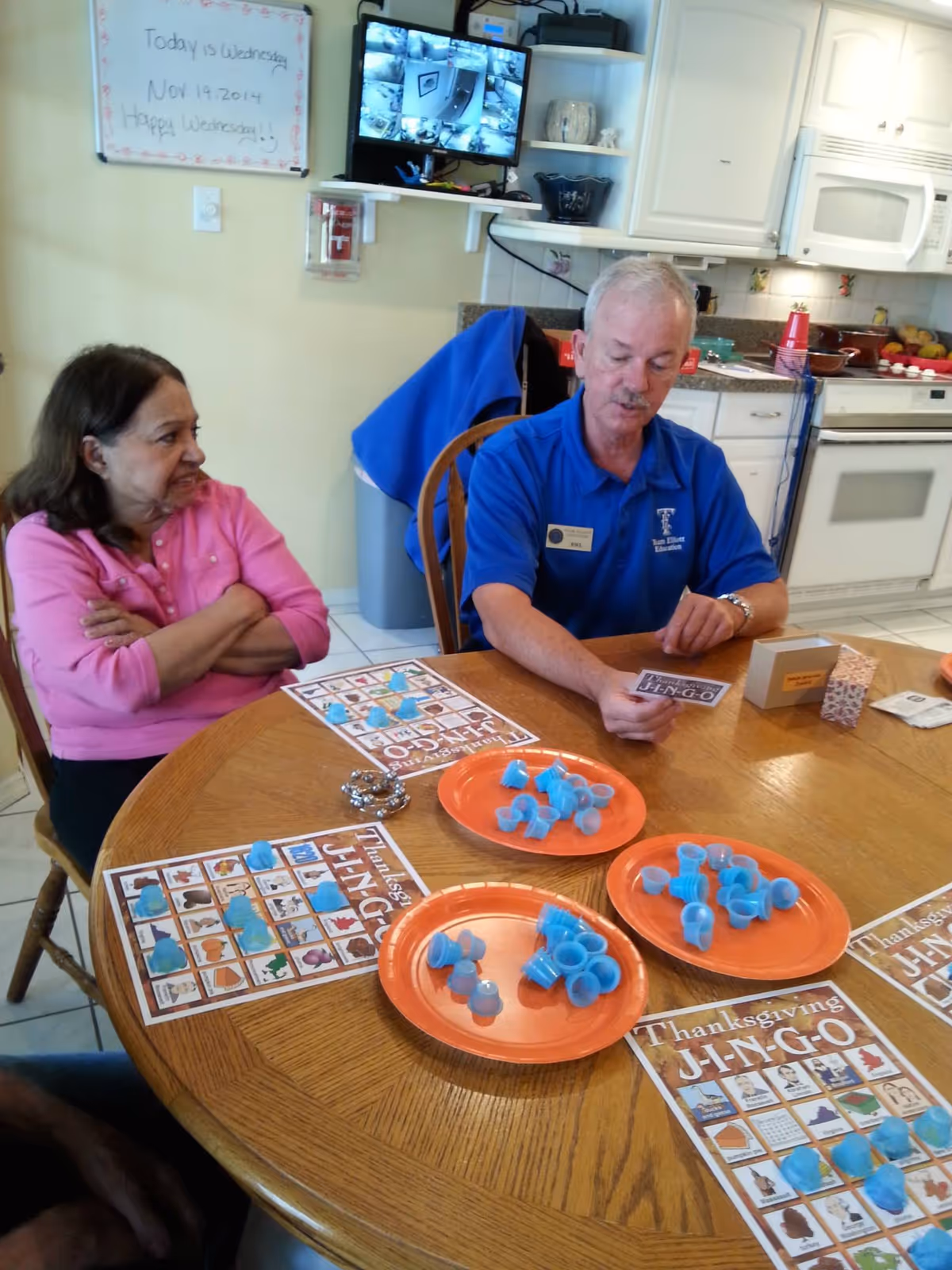 Two seniors sitting at a round table playing Thanksgiving bingo with blue markers in a communal dining/kitchen area.