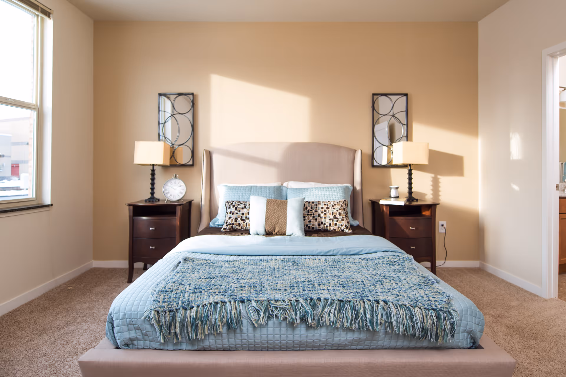 A neatly made bed with a beige upholstered headboard and light blue bedding, including a quilt and decorative pillows. On each side of the bed are dark wooden nightstands with table lamps and decorative items. Two rectangular mirrors with circular designs hang on the beige wall above the nightstands. A window is visible on the left side, and a doorway leading to another room is on the right.
