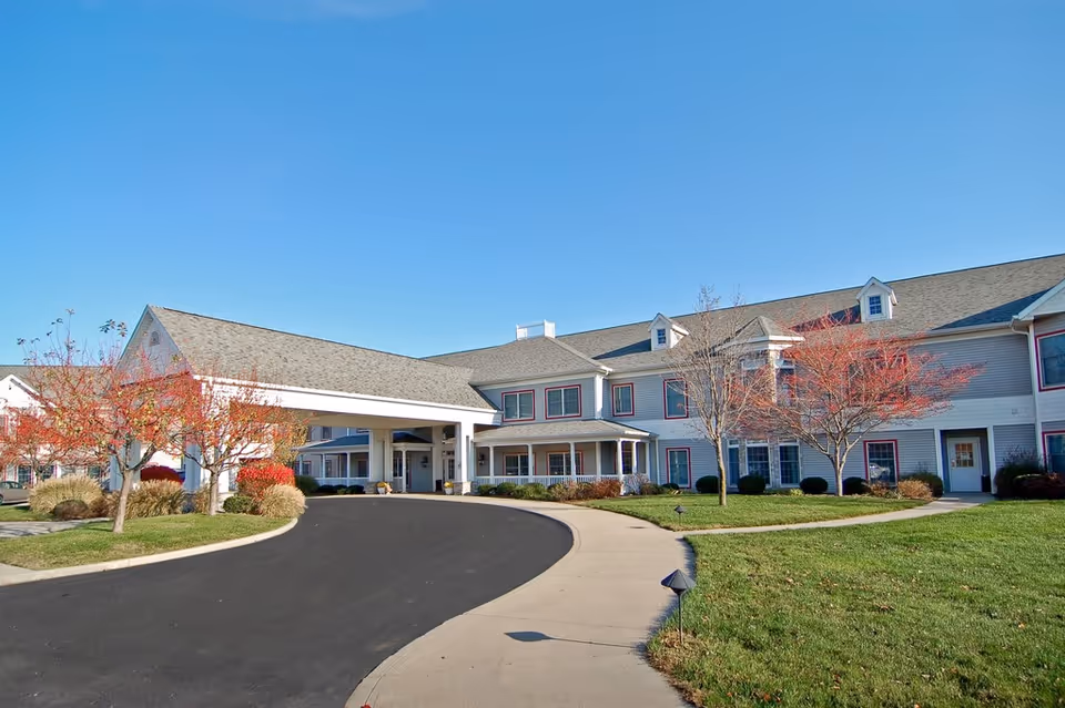 Exterior view of Wynnfield Crossing senior living facility showing a two-story building with a covered entrance, surrounded by a curved driveway, green lawn, and trees with autumn foliage under a clear blue sky.