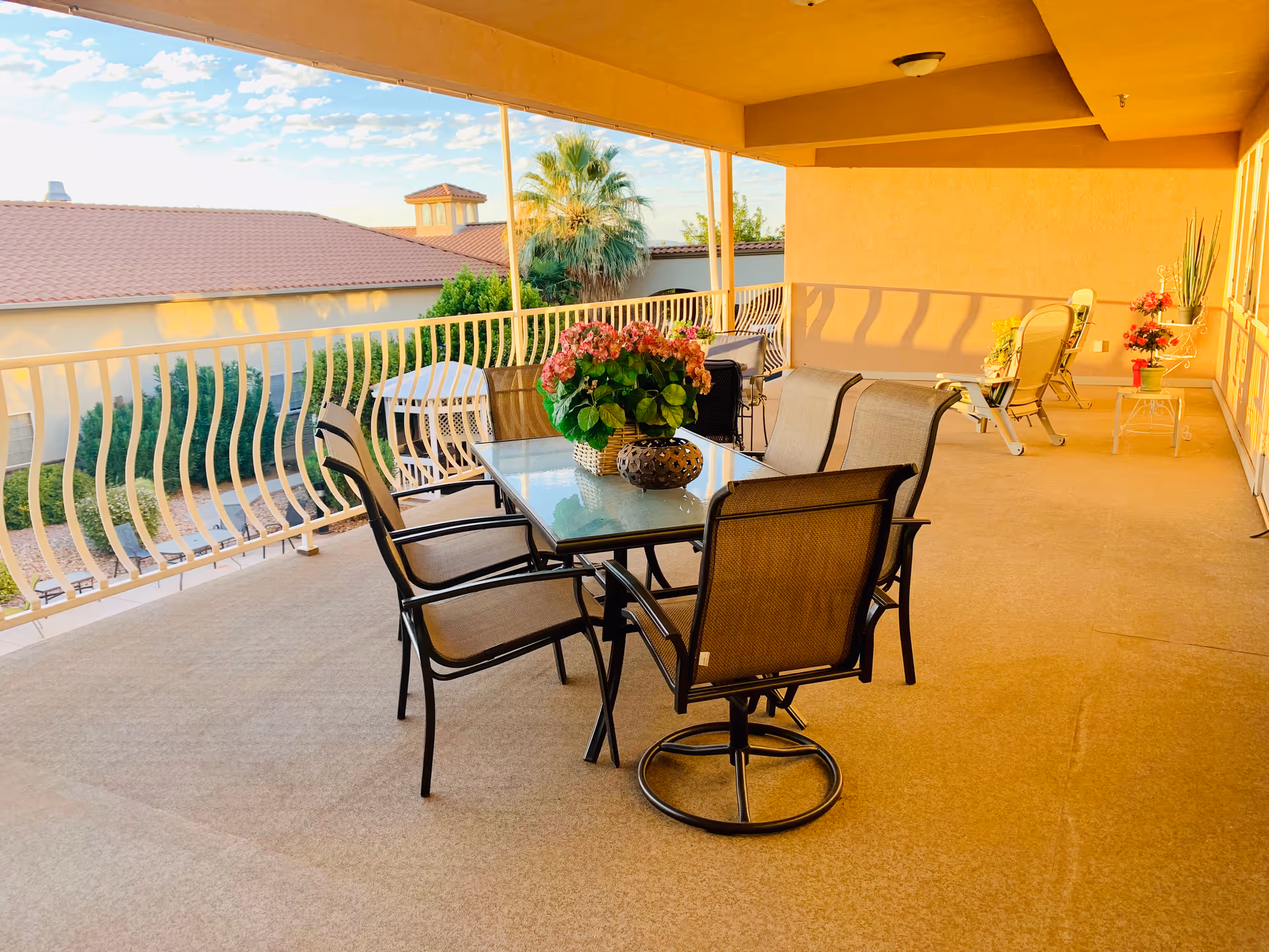 A spacious covered outdoor patio area with a glass-top table surrounded by six chairs. The table has a decorative flower arrangement and a small ornamental piece. In the background, there are additional chairs and small tables with potted plants. The patio overlooks a garden area with trees and a building with a tiled roof under a partly cloudy sky.