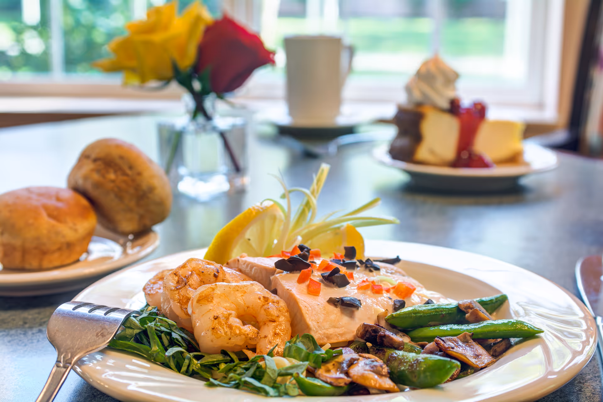 A plated meal featuring cooked shrimp, a piece of fish garnished with chopped olives and red peppers, sautéed green beans and mushrooms, and a lemon wedge on a bed of greens. In the background, there are two bread rolls on a small plate, a dessert plate with cheesecake topped with whipped cream and strawberry sauce, a white coffee cup, and a small vase with yellow and red roses on a table near a window.