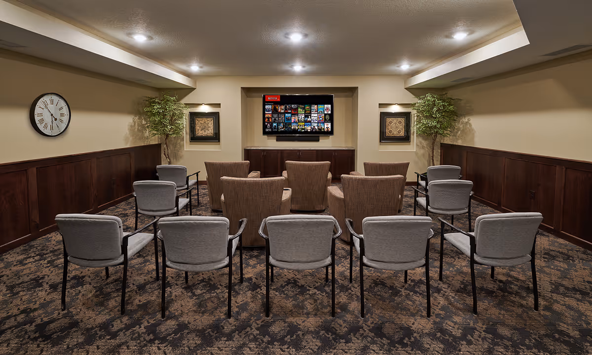 A cozy media room with rows of chairs facing a wall-mounted flat screen TV displaying a Netflix interface. The room has beige walls with dark wood paneling, two framed artworks, two potted plants, a wall clock, and recessed ceiling lights.