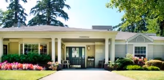 Front exterior view of Jurgens Park Senior Living building with a covered entrance, green double doors, surrounded by well-maintained landscaping including bushes, flowers, and tall trees in the background.