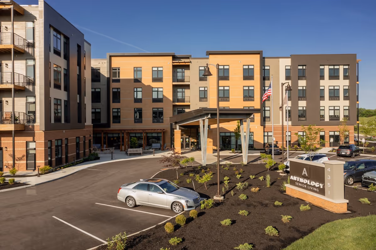 Exterior view of a modern senior living facility named Anthology Senior Living, showing a multi-story building with large windows, a covered entrance, a parking lot with several cars, landscaped greenery, and an American flag on a flagpole under a clear blue sky.