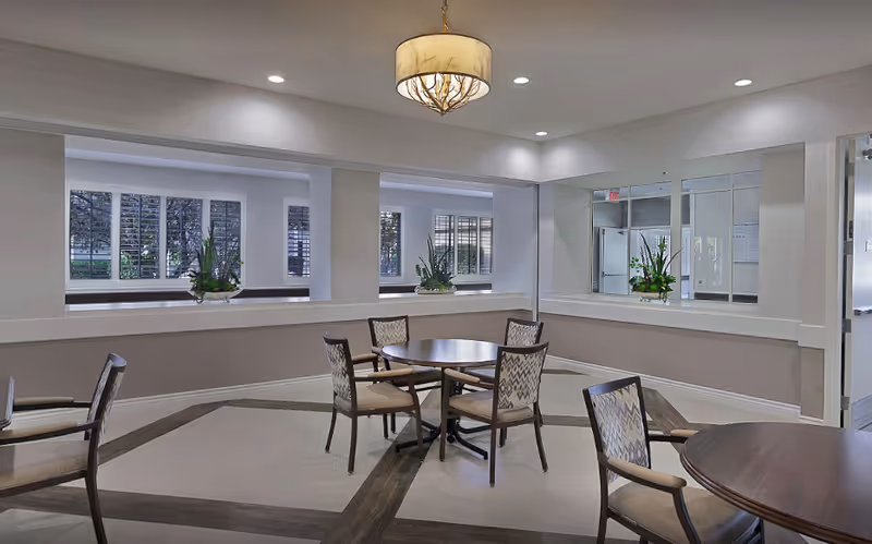 Bright communal dining area with round tables and chairs, a central chandelier, indoor plants, and interior windows to an adjacent room.
