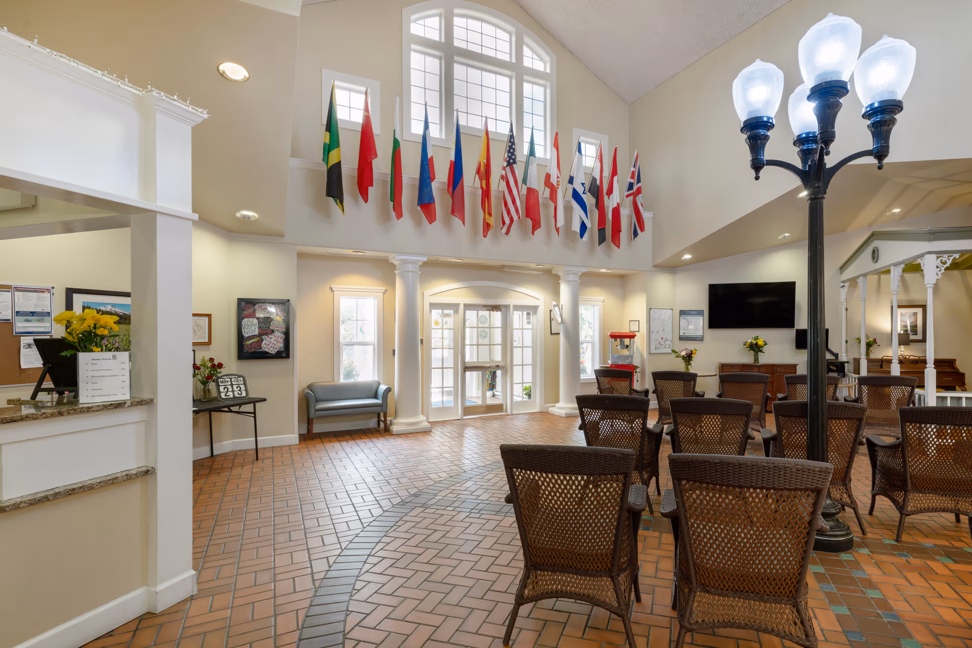 A spacious common area in a senior living facility with wicker chairs arranged facing a large flat-screen TV mounted on the wall. The room features a high ceiling with large windows and multiple international flags hanging above the entrance. There is a decorative streetlamp-style light fixture in the center, a popcorn machine near the entrance, and a piano in the corner. The floor is tiled with a brick pattern, and there are columns framing the entrance doors.