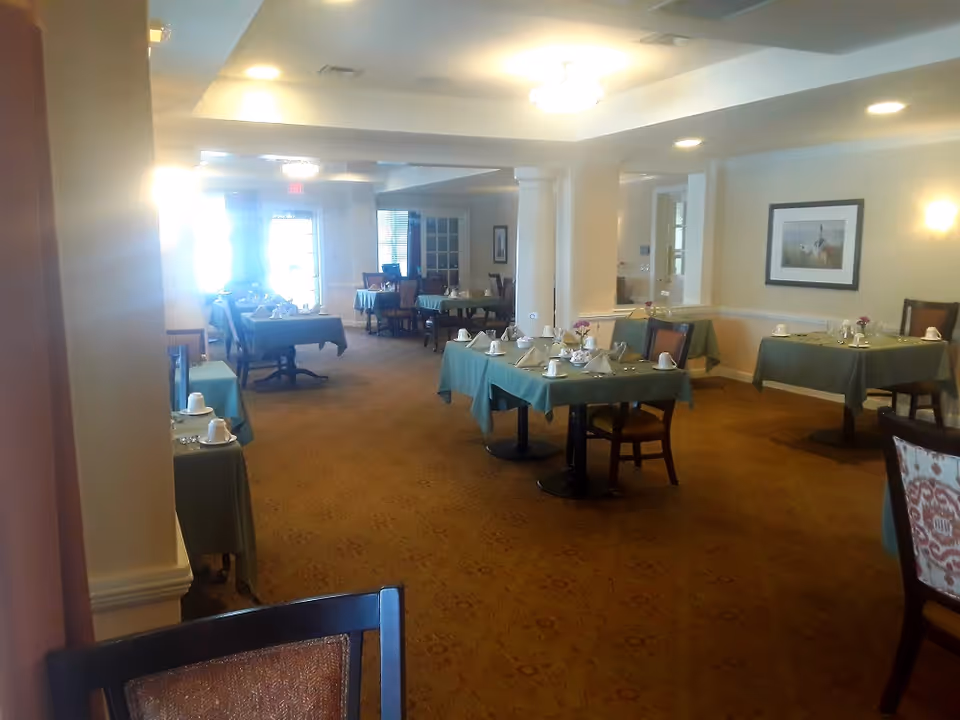 Dining room with multiple small tables covered in green tablecloths set for a meal in a senior living facility.