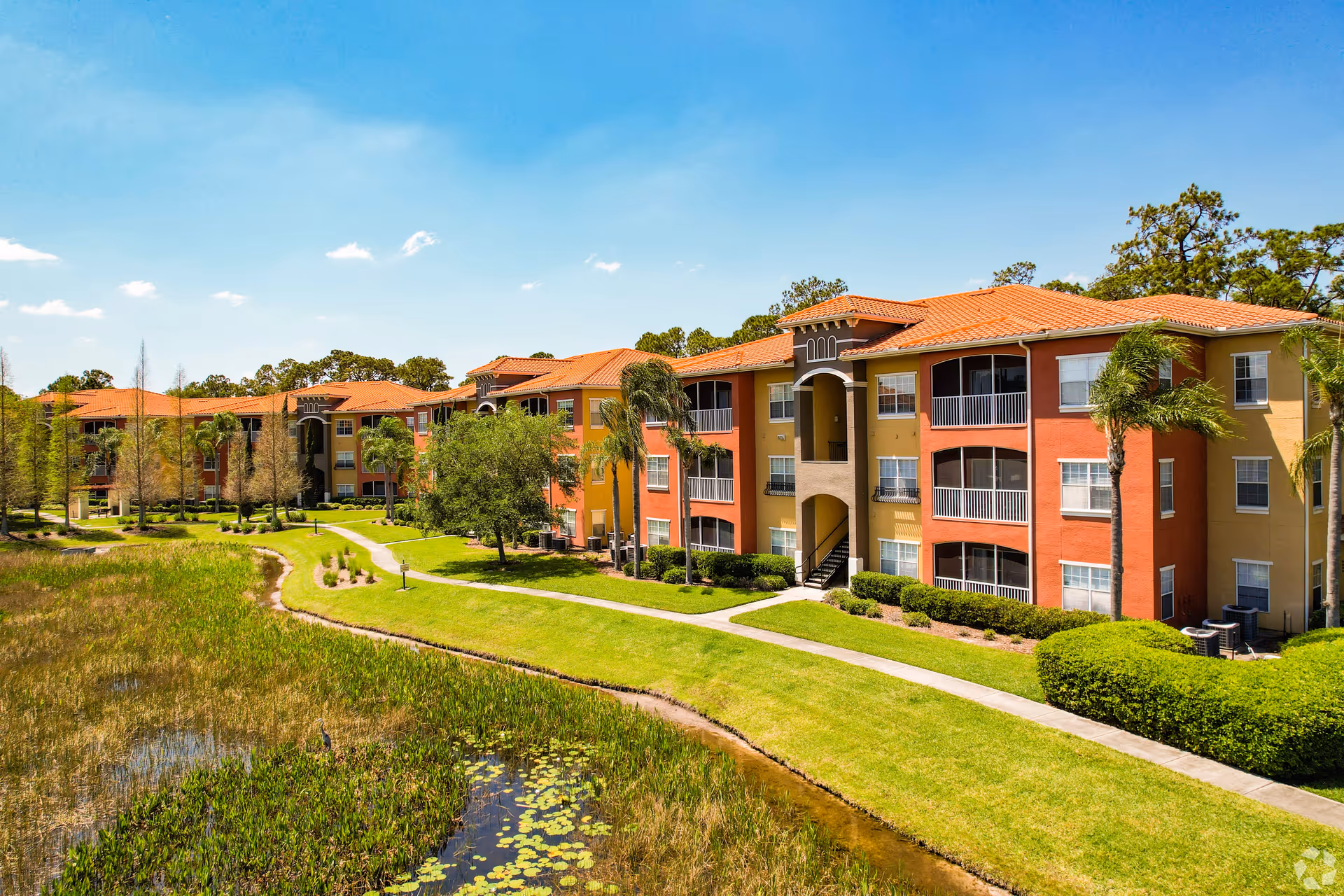 Exterior view of a multi-story residential building with orange and yellow walls and red tile roofs, surrounded by green lawns, palm trees, and a small pond with water plants under a clear blue sky.