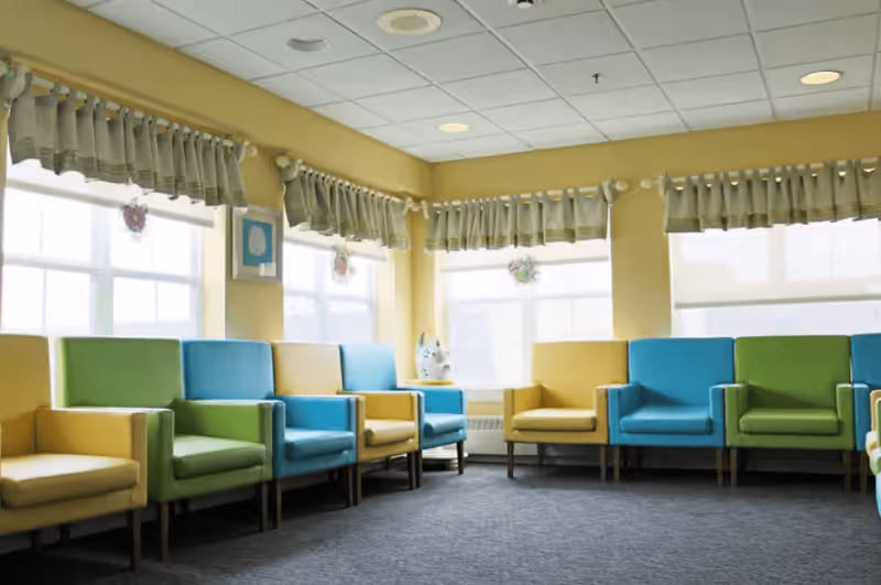 A bright room with large windows covered by short curtains, featuring a row of colorful armchairs in yellow, green, and blue arranged along the walls on a carpeted floor.