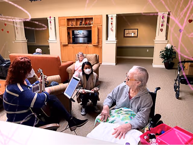 Residents and staff gather in a communal living room where a musician plays guitar, a masked caregiver kneels beside a woman in a wheelchair, and another resident sits on a couch.