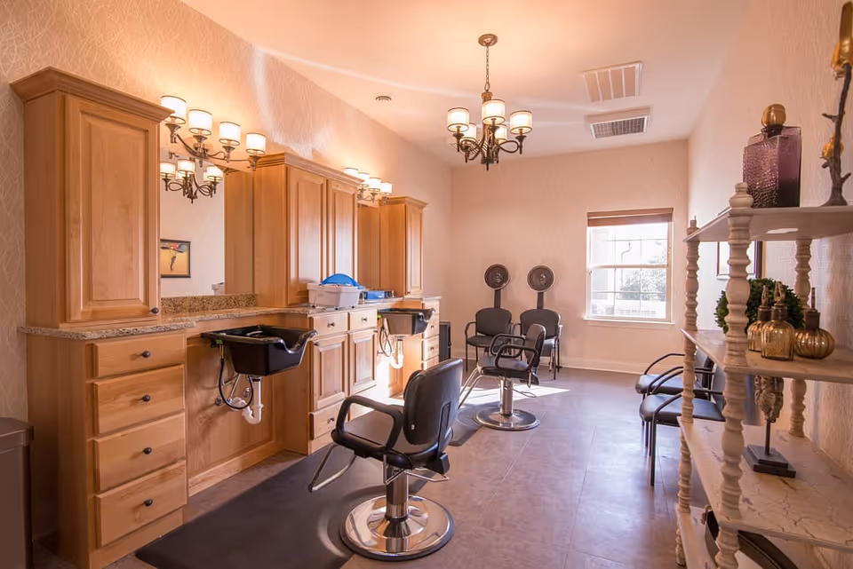 A well-lit salon room with wooden cabinetry, two black salon chairs in front of sinks, and two hair dryer chairs near a window. The room has beige walls, a chandelier, wall-mounted lights, and a shelving unit with decorative items.