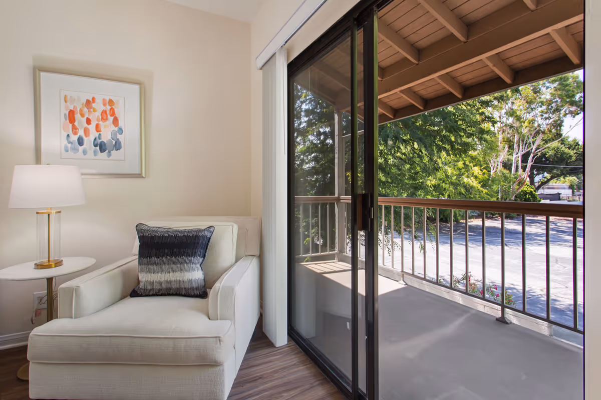 A cozy corner of a living room featuring a white armchair with a decorative pillow, a small round side table with a lamp, and a framed abstract painting on the wall. Next to the chair is a large sliding glass door leading to a covered balcony with a view of trees and a parking area outside.