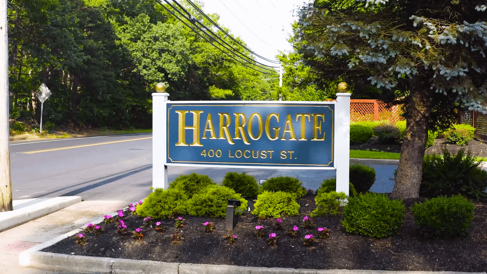 Outdoor view of a sign for Harrogate Retirement Community at 400 Locust St, surrounded by landscaped bushes and flowers with a road and trees in the background.