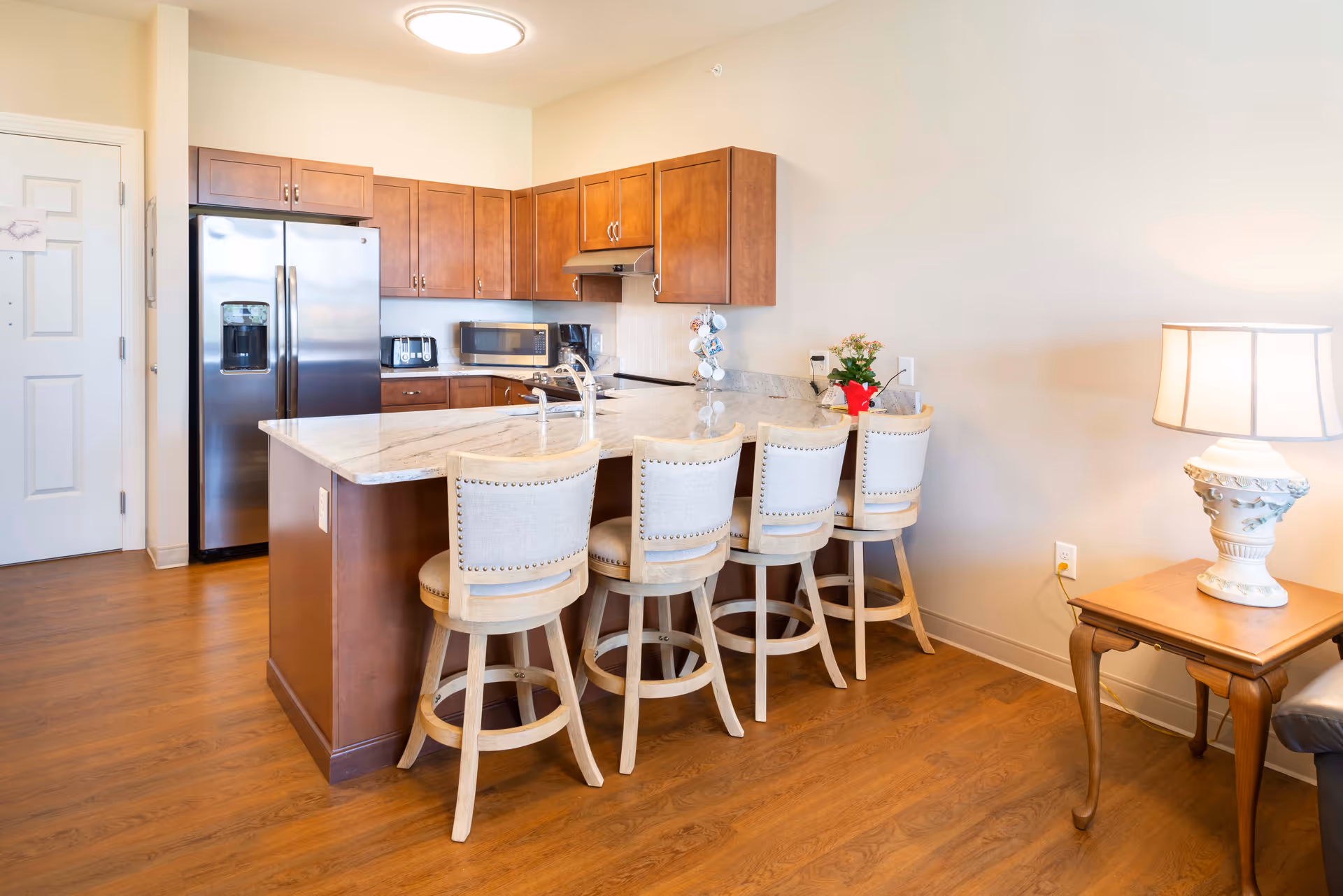 A bright kitchen area with wooden cabinets, a stainless steel refrigerator, microwave, toaster, coffee maker, and a large marble countertop island with four cushioned bar stools. To the right, there is a wooden side table with a decorative lamp and a small potted plant on the countertop.