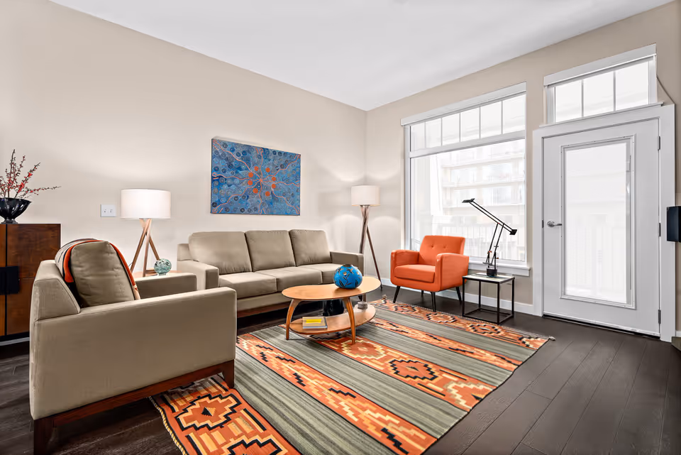 A bright living room with beige walls and dark wooden floors. The room features a beige sofa and armchair, an orange armchair, a wooden coffee table with a blue decorative ball, and a colorful patterned rug. There are two floor lamps, a piece of abstract art on the wall, a side table with a black lamp, and large windows letting in natural light. A white door with a window is also visible.