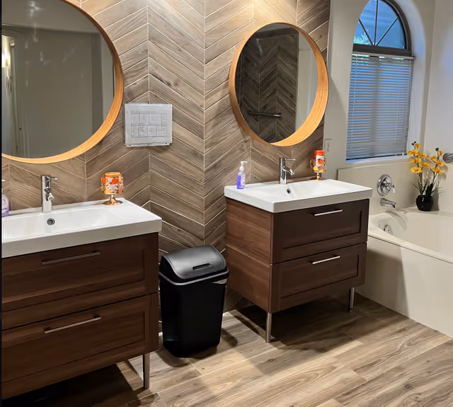 Modern bathroom with two wood vanities and round mirrors, a bathtub by a window, chevron-tiled wall, and a trash bin.