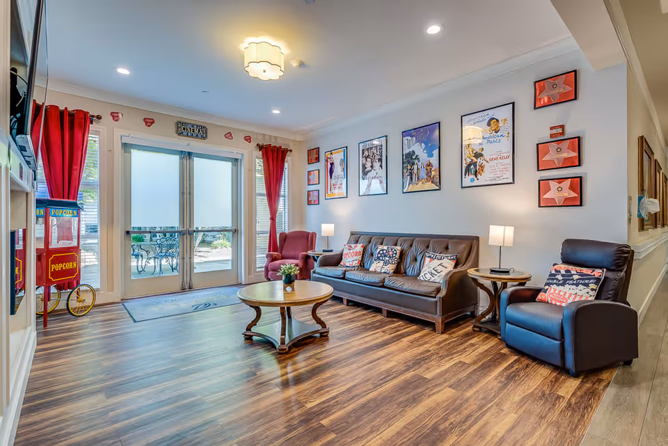 A cozy senior living facility lounge area with a brown leather sofa, two armchairs, a round wooden coffee table, and side tables with lamps. The walls are decorated with vintage movie posters and framed star plaques. There is a popcorn machine near glass double doors with red curtains, leading to an outdoor patio with metal chairs and tables.