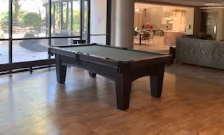 Interior view of a common area in a senior living facility featuring a pool table on a wooden floor near large glass windows with a view of outdoor greenery. In the background, there is a seating area with tables and chairs and a counter or reception desk.