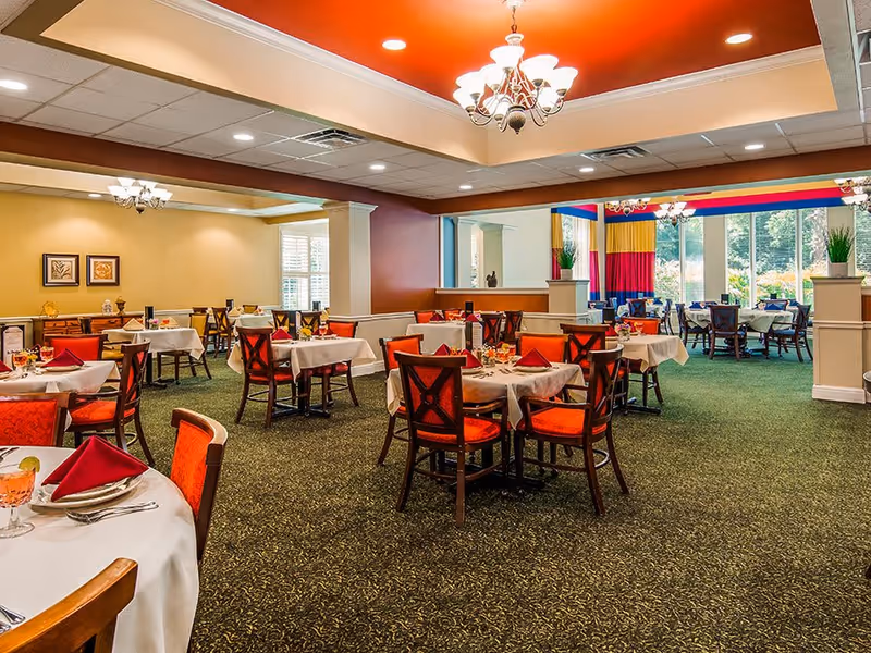 A spacious dining room with multiple tables covered in white tablecloths and set with plates, glasses, and red napkins. The chairs have red cushions and dark wooden frames. The room features a green carpet, yellow and red walls, large windows letting in natural light, and chandeliers hanging from the ceiling.