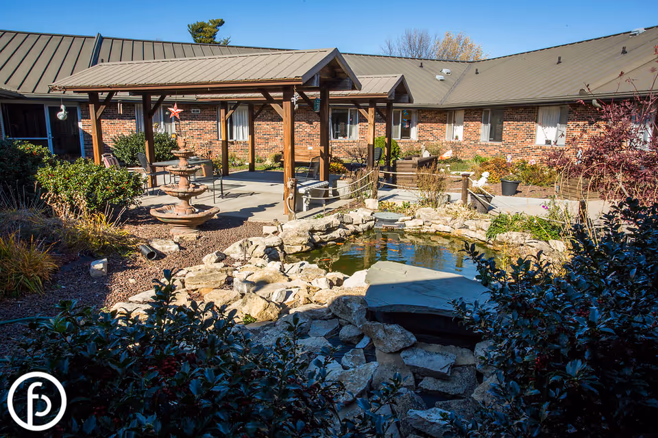 Sunny courtyard with a wooden pergola, outdoor seating, a tiered fountain and rock-lined pond in front of a single-story brick building.