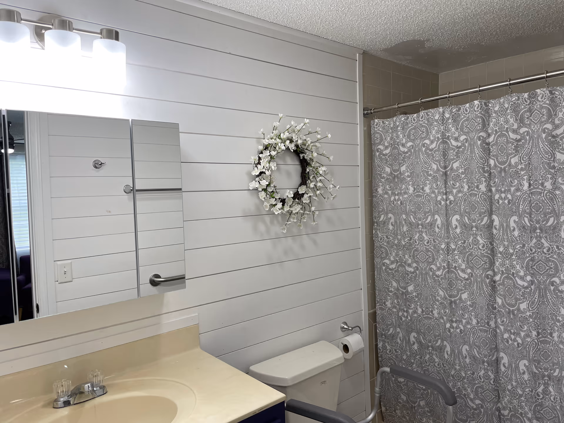 A bathroom with a beige countertop and sink, a mirror with three panels above the sink, a toilet with a grab bar, a toilet paper holder, a decorative white floral wreath on the wall, and a shower with a gray and white patterned curtain.