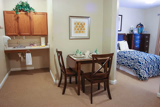 A cozy senior living apartment featuring a small dining area with a wooden table set for two, a kitchenette with wooden cabinets and a sink, and a bedroom area with a bed covered in a blue and white patterned comforter, a dresser, and framed artwork on the walls.