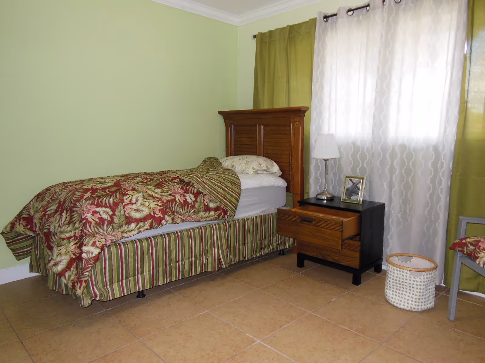 A bedroom with a single bed featuring a wooden headboard, floral and striped bedding, a nightstand with a lamp and a framed photo, green curtains, and a woven basket on a tiled floor.