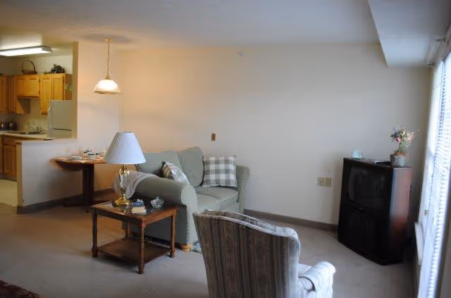 A cozy living room with a sofa, armchair, coffee table, small corner TV, and a view into the adjacent kitchen.