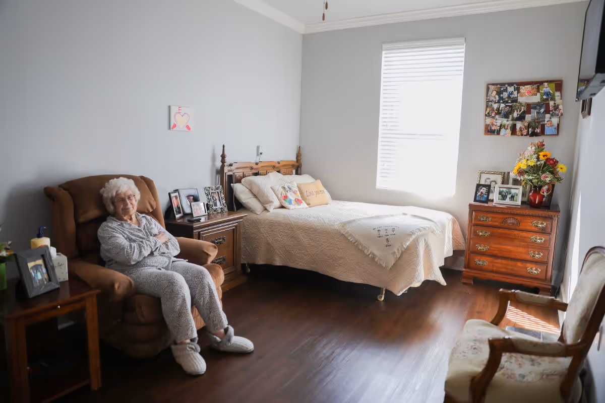 An elderly woman sitting in a brown recliner chair in a cozy bedroom with a bed, wooden nightstand, dresser with flowers and framed photos, and a window with blinds letting in natural light.