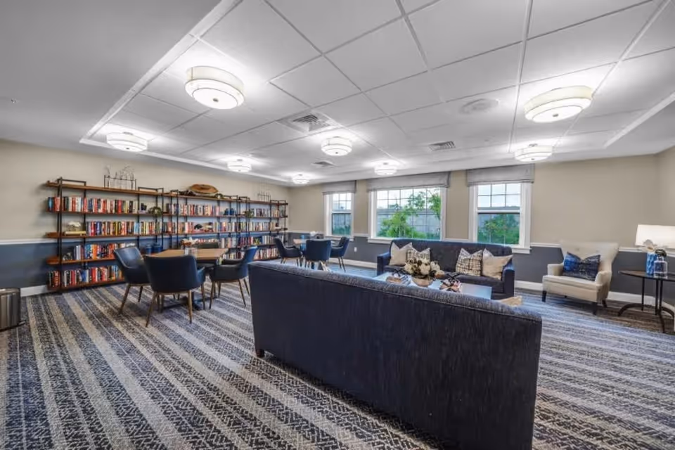 A spacious and well-lit common area with blue sofas, armchairs, and tables. There is a large bookshelf filled with books along one wall. The room has large windows letting in natural light and a patterned carpet covering the floor.