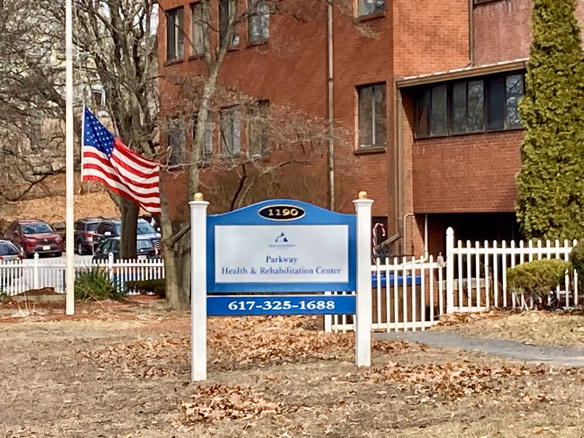 Outdoor view of Parkway Health & Rehabilitation Center sign with the address number 1190 on top. The sign is blue and white with a phone number 617-325-1688 displayed. Behind the sign is a white picket fence, some trees, and a red brick building. An American flag is flying on a flagpole to the left of the sign.
