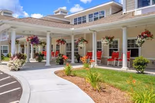Exterior view of a senior living facility with a covered porch featuring white columns, hanging flower baskets, red rocking chairs, and landscaped garden beds under a blue sky with some clouds.