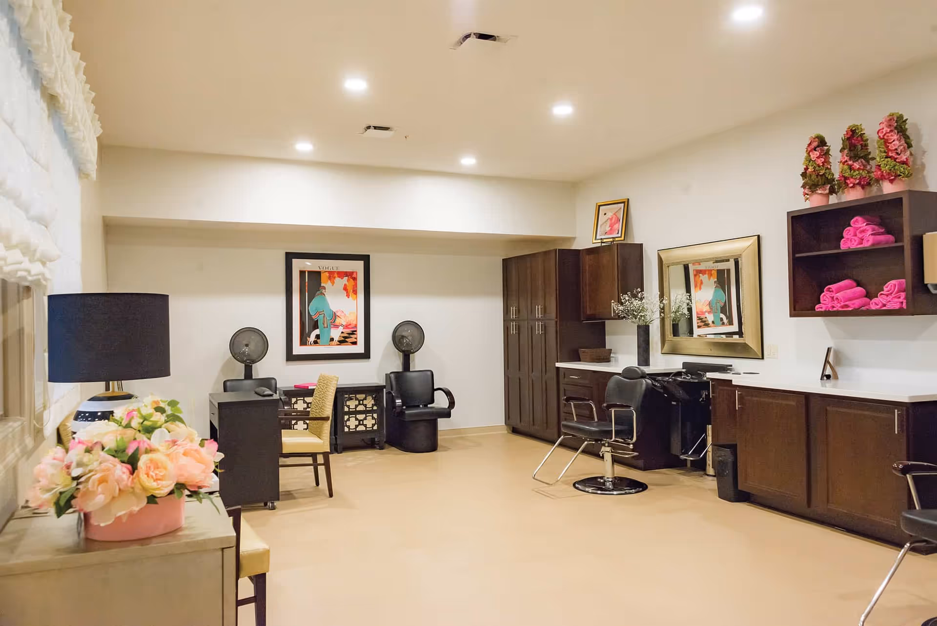 A well-lit salon room with beige flooring and white walls. The room features dark wooden cabinets and shelves with neatly rolled pink towels. There are two black salon chairs, a large mirror above a counter, and two hair dryer stations against the back wall. Decorative elements include framed artwork, a vase with flowers, and a lamp with a black shade on a side table with a flower arrangement.